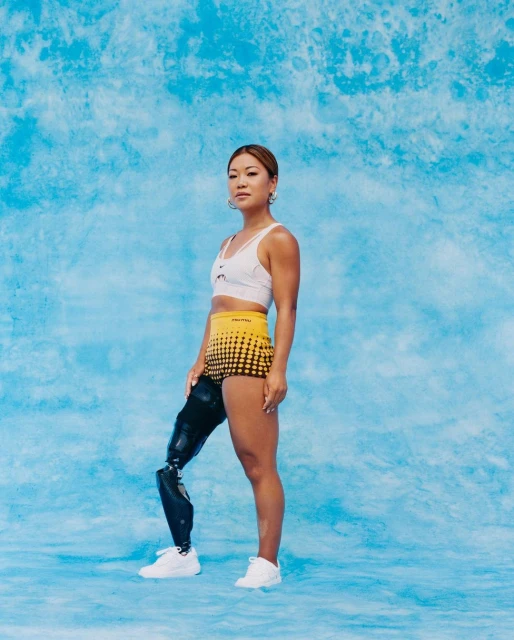 Young woman with prosthetic leg poses confidently in athletic wear against a marbled light blue backdrop in a studio shot.