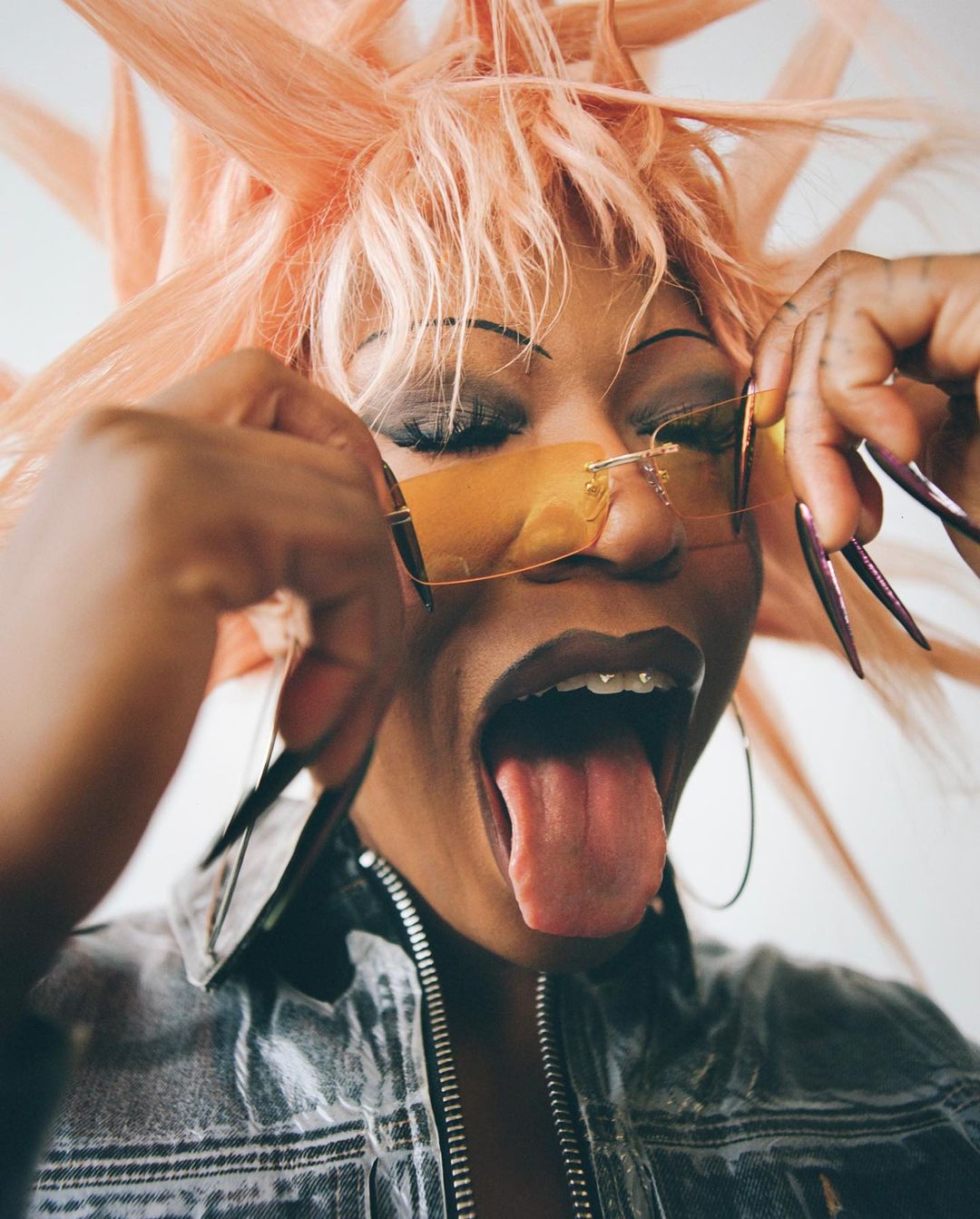 Close-up portrait of a woman with a peach wig, sunglasses, and a denim jacket, sticking her tongue out in front of a white background.