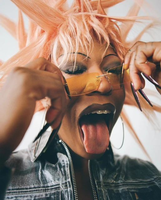 Close-up portrait of a woman with a peach wig, sunglasses, and a denim jacket, sticking her tongue out in front of a white background.