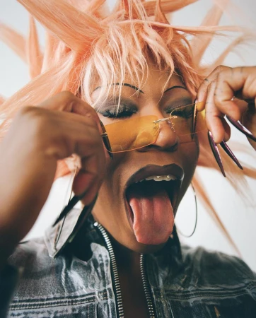 Close-up portrait of a woman with a peach wig, sunglasses, and a denim jacket, sticking her tongue out in front of a white background.