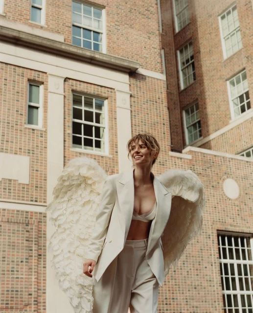 Smiling woman in a white suit and lingerie with feathered angel wings stands in front of a brick building on a sunny day.