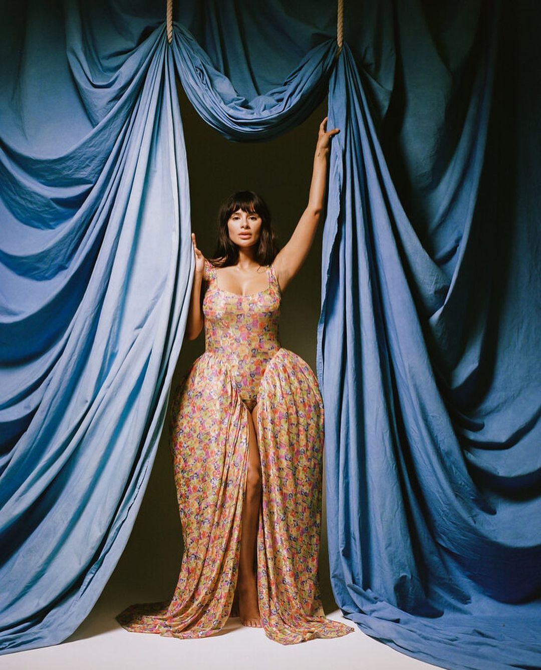 Portrait of an adult woman in a floral dress standing between blue drapes, looking at the camera with a neutral expression.