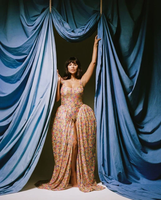 Portrait of an adult woman in a floral dress standing between blue drapes, looking at the camera with a neutral expression.