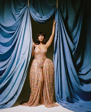 Portrait of an adult woman in a floral dress standing between blue drapes, looking at the camera with a neutral expression.