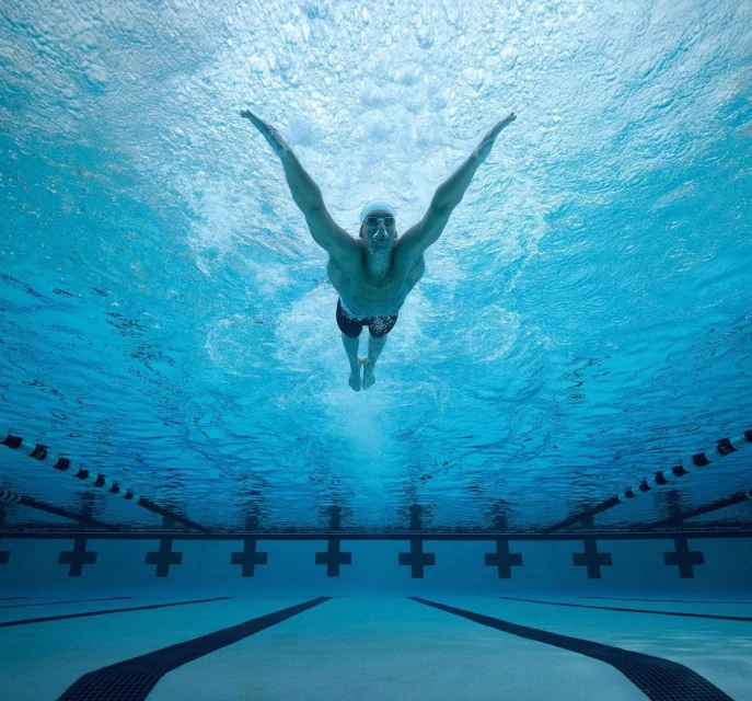 Underwater shot of an adult male swimmer with arms outstretched, wearing goggles and a swim cap, in an indoor pool with lane markings.