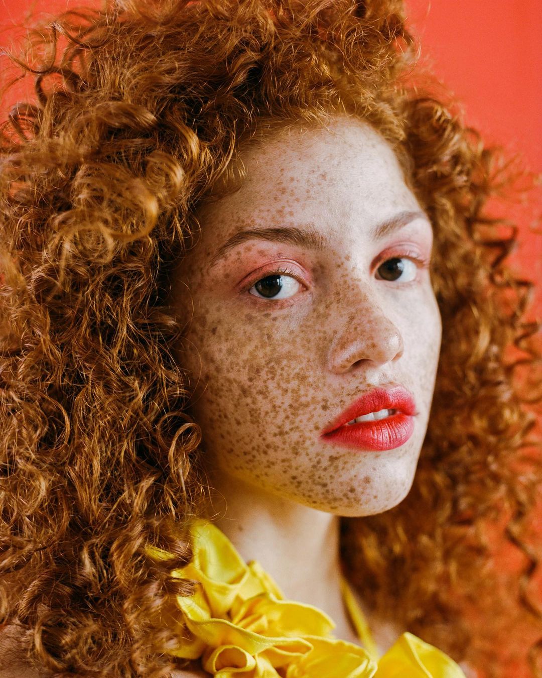 Close-up portrait of a woman with red curly hair, freckles, red lipstick, and a yellow ruffled collar against an orange background.