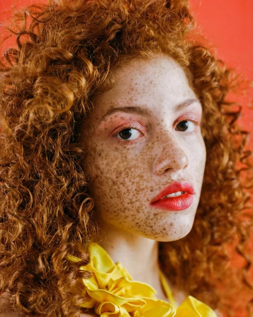 Close-up portrait of a woman with red curly hair, freckles, red lipstick, and a yellow ruffled collar against an orange background.