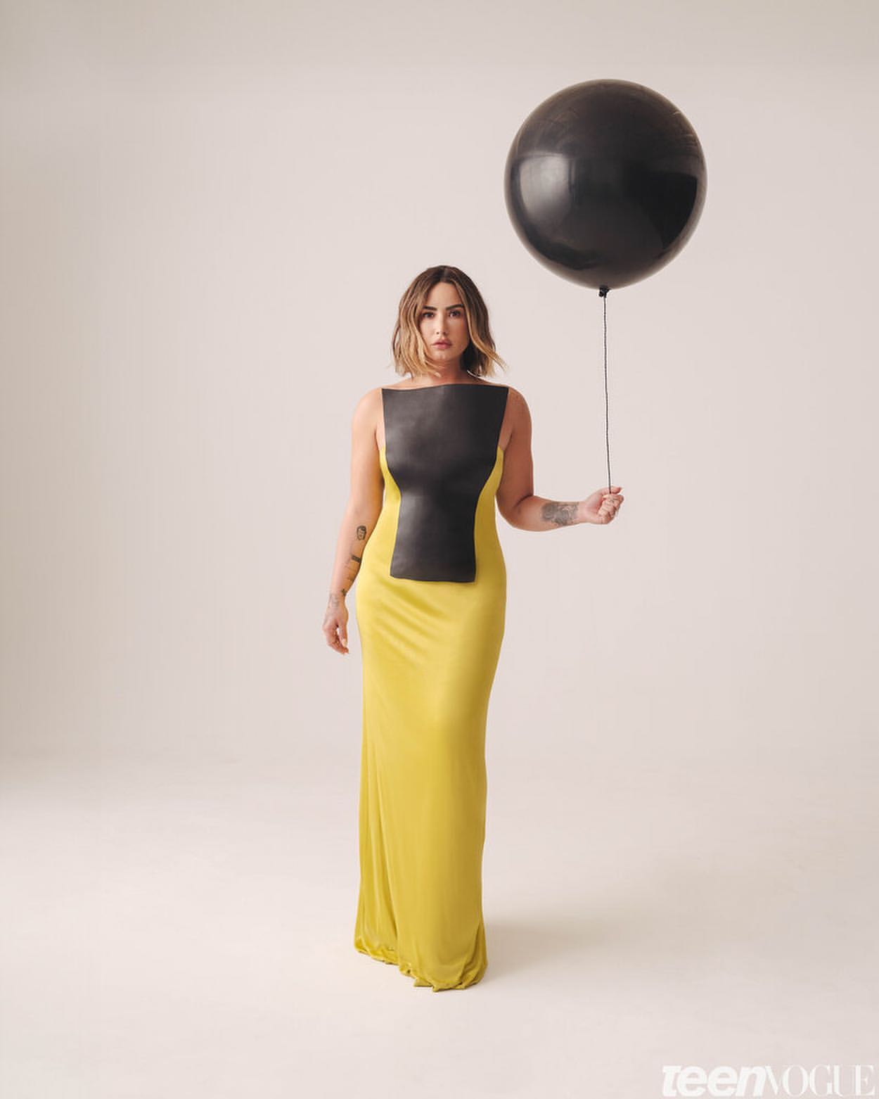Adult woman posing in a yellow dress with a black top holds a large black balloon on a neutral background in a studio portrait.