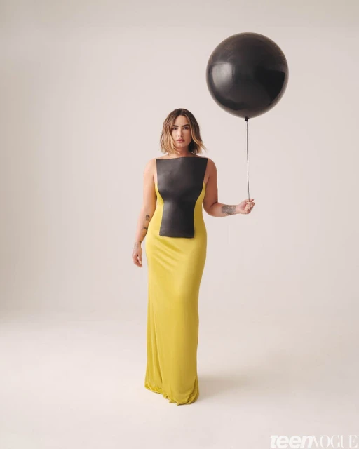 Adult woman posing in a yellow dress with a black top holds a large black balloon on a neutral background in a studio portrait.