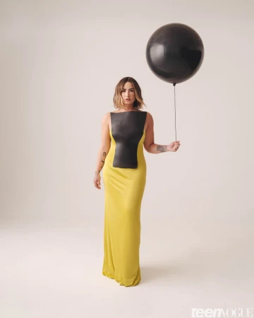 Adult woman posing in a yellow dress with a black top holds a large black balloon on a neutral background in a studio portrait.