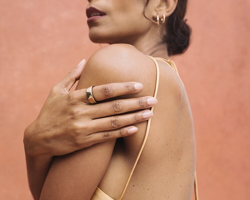 Woman with tan skin poses with a gold ring and hoop earrings against a textured orange background in a beauty or fashion context.