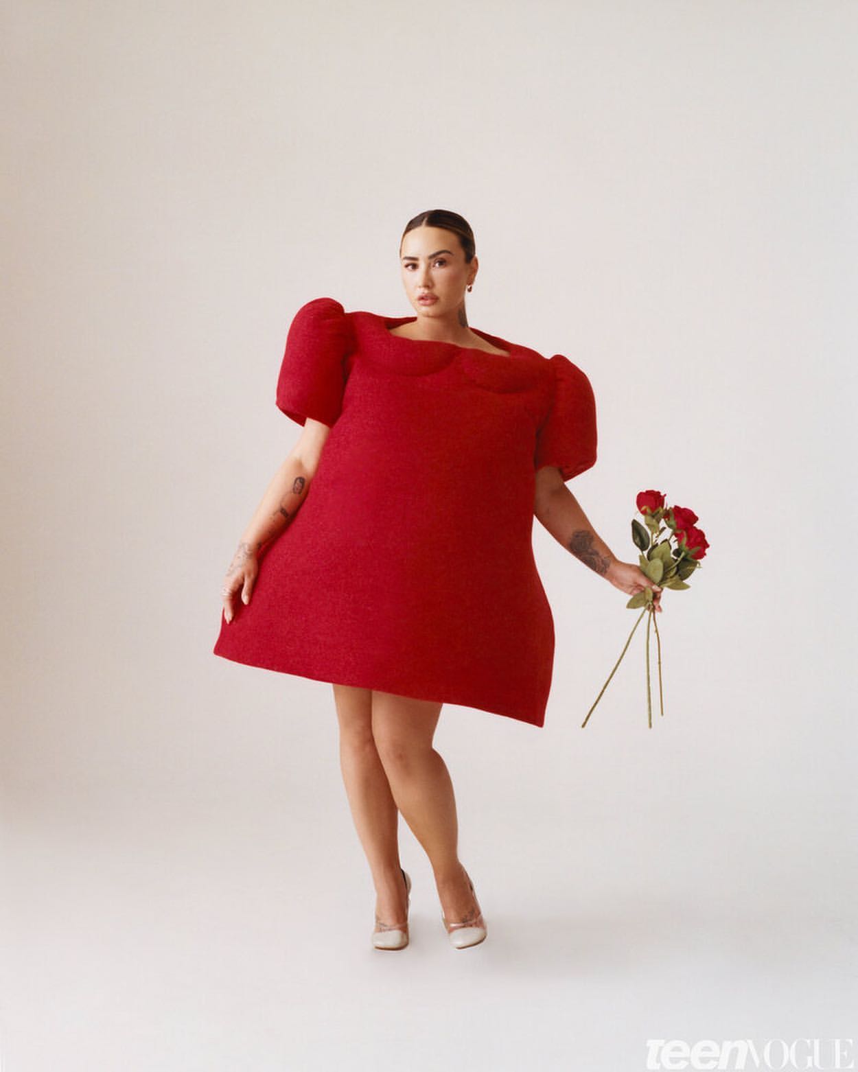 A stylish woman in a red dress and heels holds roses against a plain background, offering a striking visual for fashion and beauty.