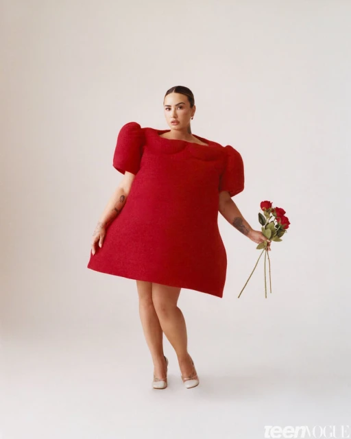 A stylish woman in a red dress and heels holds roses against a plain background, offering a striking visual for fashion and beauty.