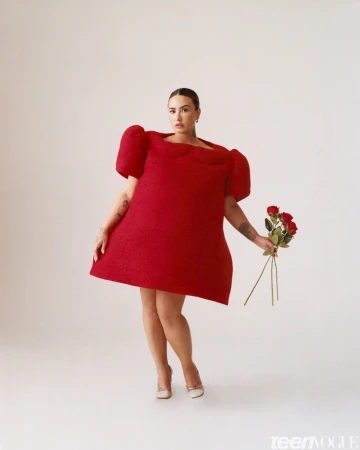 A stylish woman in a red dress and heels holds roses against a plain background, offering a striking visual for fashion and beauty.