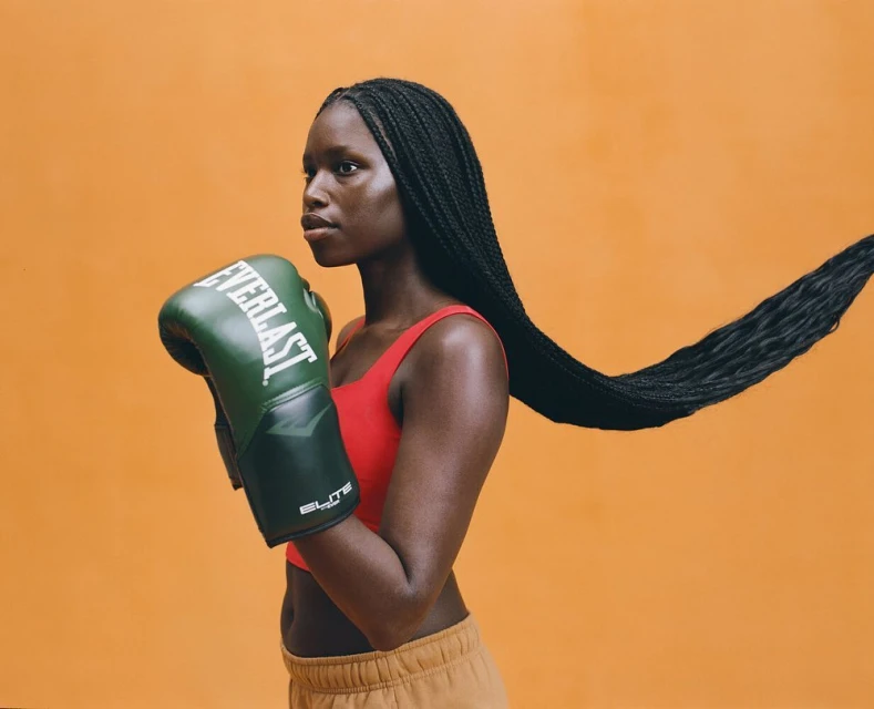 African-American woman in sports bra with boxing gloves. Athletic female boxer poses in a studio with braids against an orange backdrop.