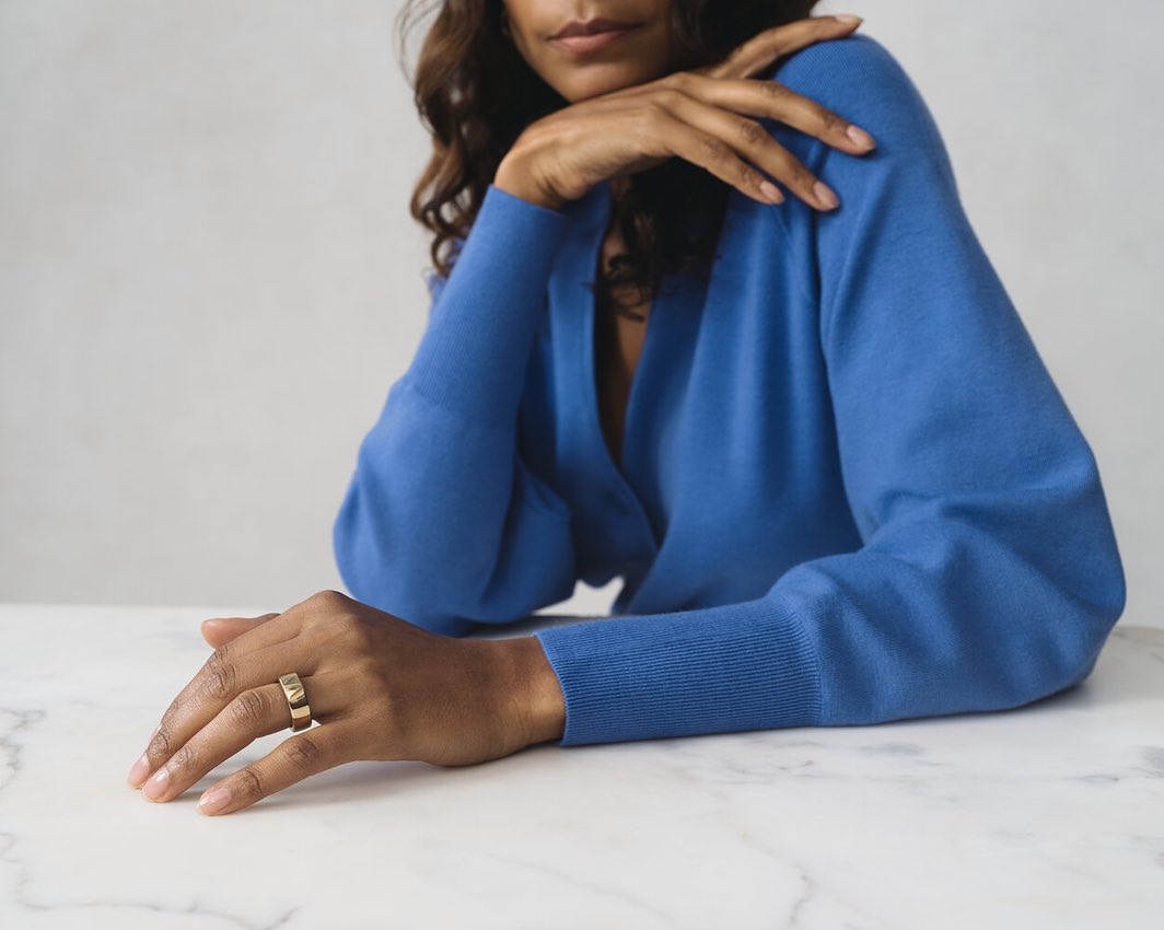 A woman rests her hands on a marble table, showcasing a gold ring on her finger, wearing a blue sweater in a softly lit, neutral setting.