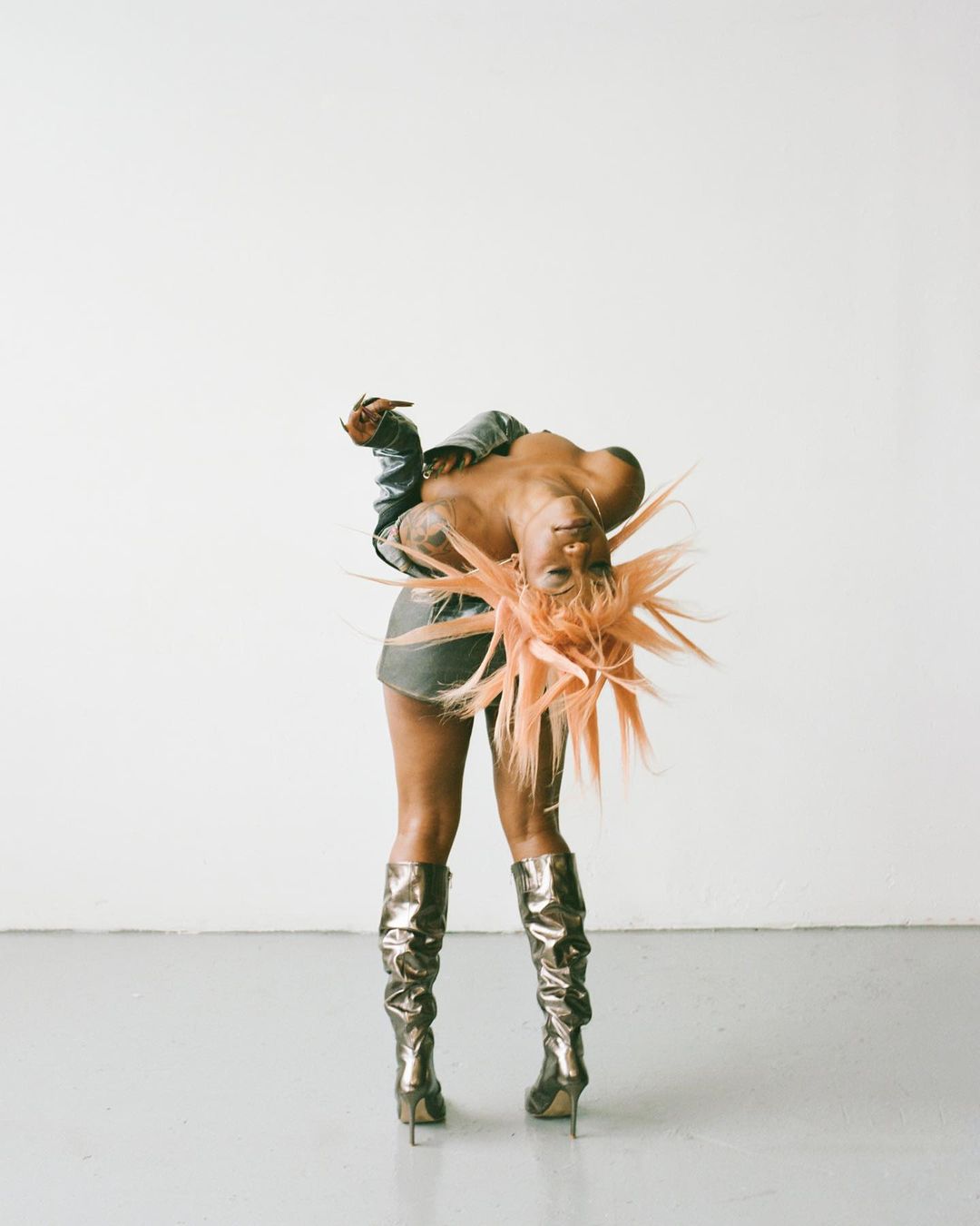 Woman in metallic boots and a denim jacket poses in a studio against a neutral background with an orange spiky wig.