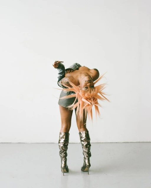 Woman in metallic boots and a denim jacket poses in a studio against a neutral background with an orange spiky wig.