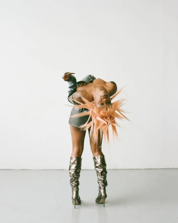 Woman in metallic boots and a denim jacket poses in a studio against a neutral background with an orange spiky wig.
