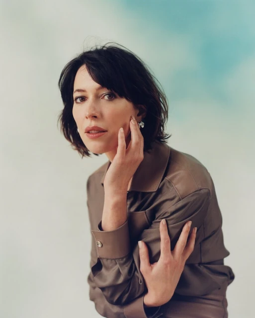 Portrait of a woman with dark hair and a brown shirt, posing with her hand near her face against a blue and white cloud background.