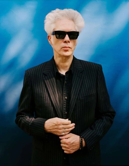 Portrait of a middle-aged adult male with silver hair and sunglasses wearing a striped suit against a blue patterned background.