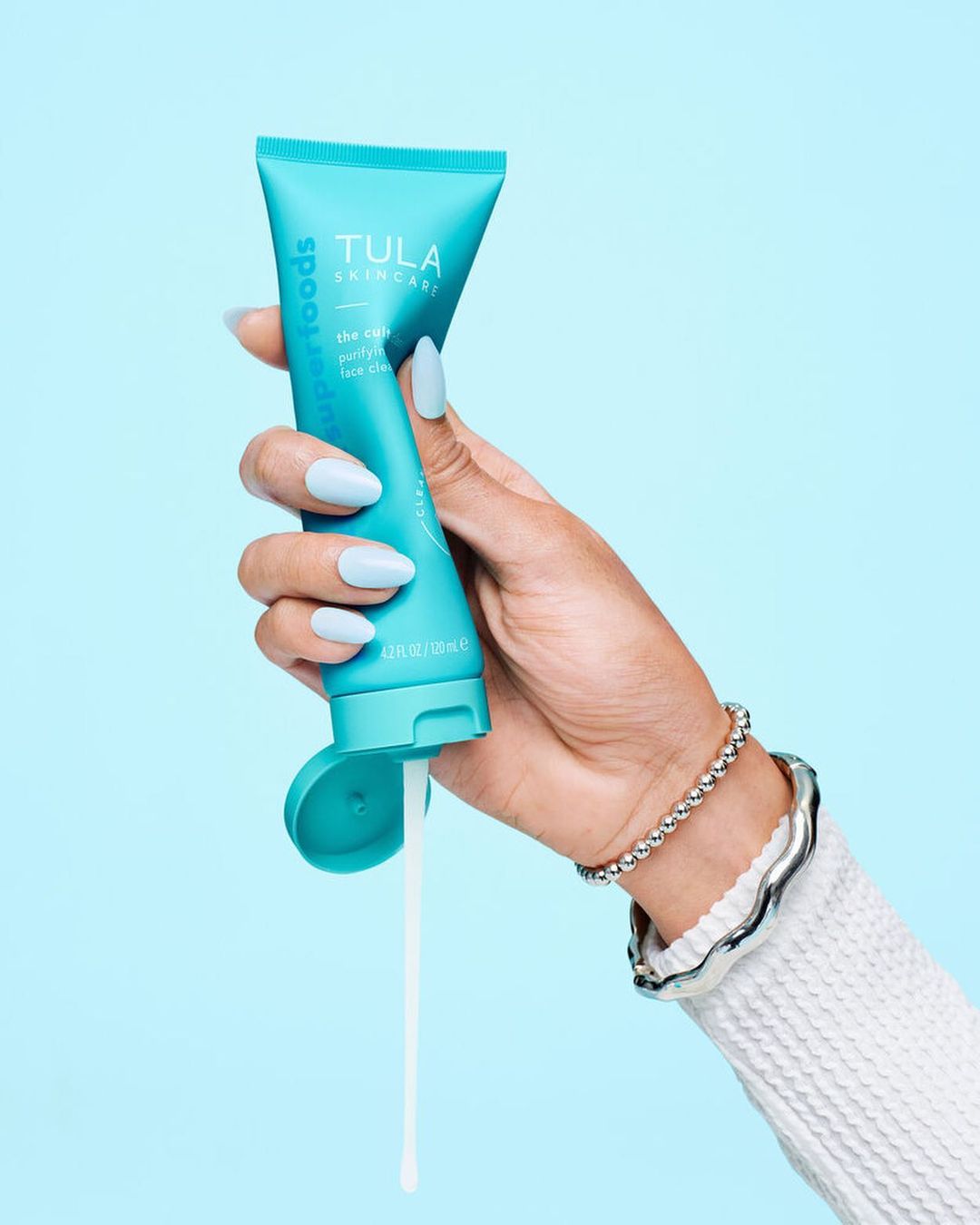 Hand holding a tube of Tula skincare product, teal packaging, white lotion, soft blue background. Beauty and skincare photography.
