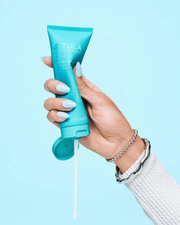 Hand holding a tube of Tula skincare product, teal packaging, white lotion, soft blue background. Beauty and skincare photography.