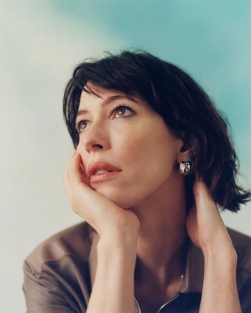 Portrait of a woman with dark hair resting her chin on her hands, wearing statement earrings against a soft background.