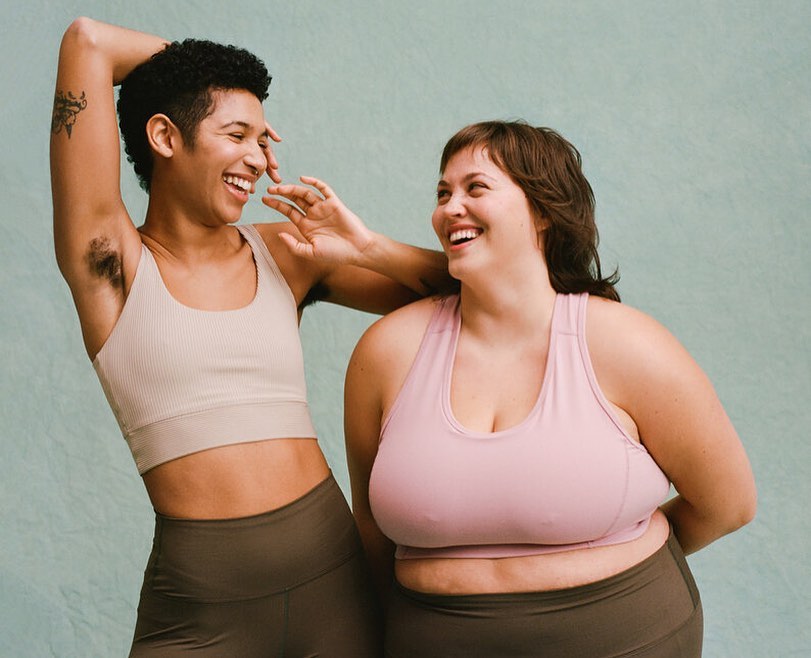 Two smiling women in sports bras and leggings pose indoors against a plain backdrop. One woman raises an arm, laughing.