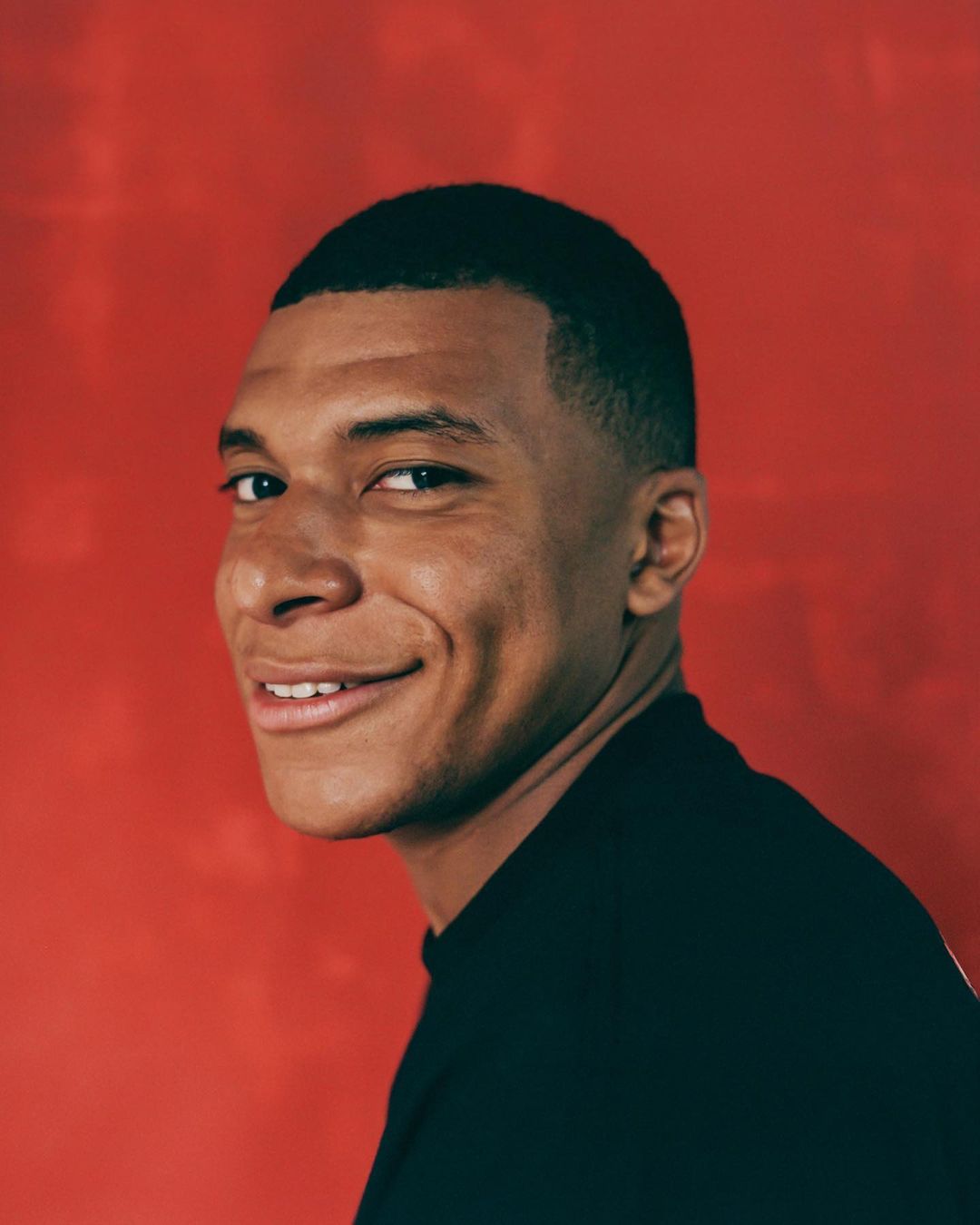 Portrait of Kylian Mbappe smiling slightly against a textured red background, photographed by Josefina Santos.