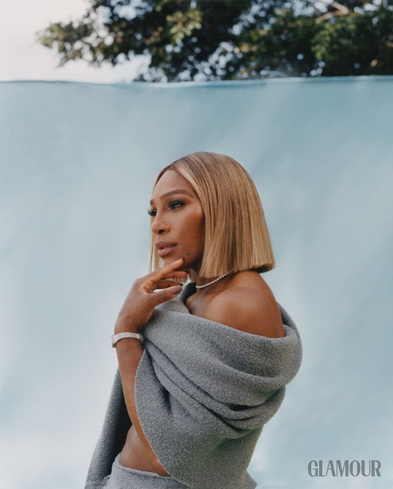 Glamour portrait of an adult woman with blonde hair and a grey sweater wrapped around her shoulders, posing in front of a pale blue background.