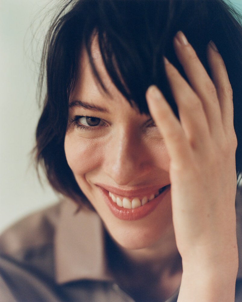 Close-up portrait of a woman with dark hair smiling while holding her hand up to her forehead.