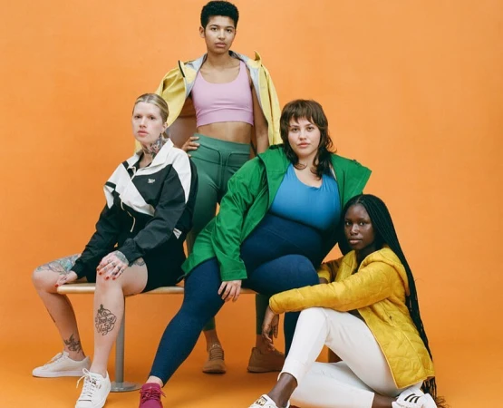Group of four diverse women in athletic wear pose in a studio setting with orange background. They are wearing a variety of colorful jackets.