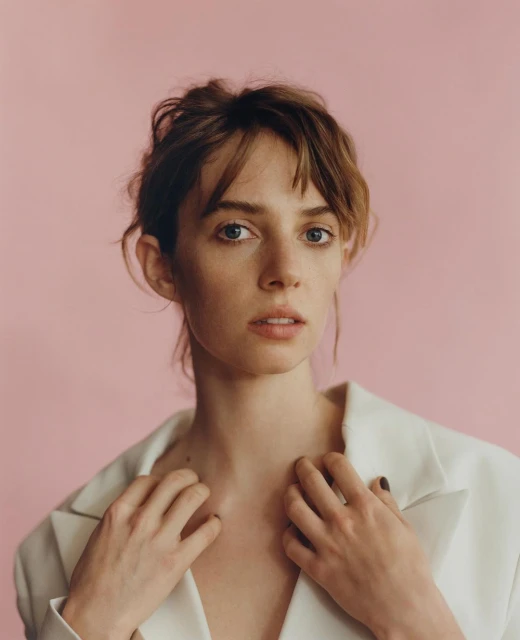 Close-up portrait of a young woman with fair skin and brown hair wearing a white jacket against a pink background in a photo studio.