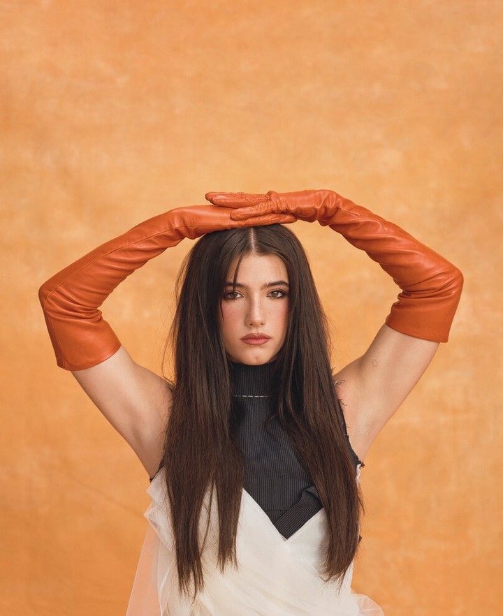 Brunette woman in studio portrait wearing white dress and orange gloves posing against orange background with neutral facial expression.