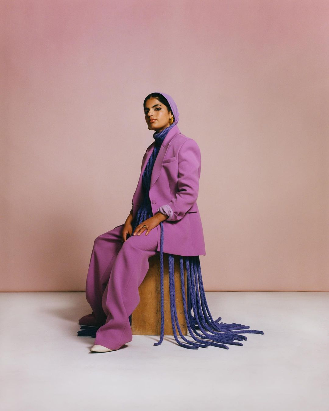 Portrait of an adult woman in a lilac pantsuit and headscarf, seated on a wooden cube against a blush-colored background in a studio setting.