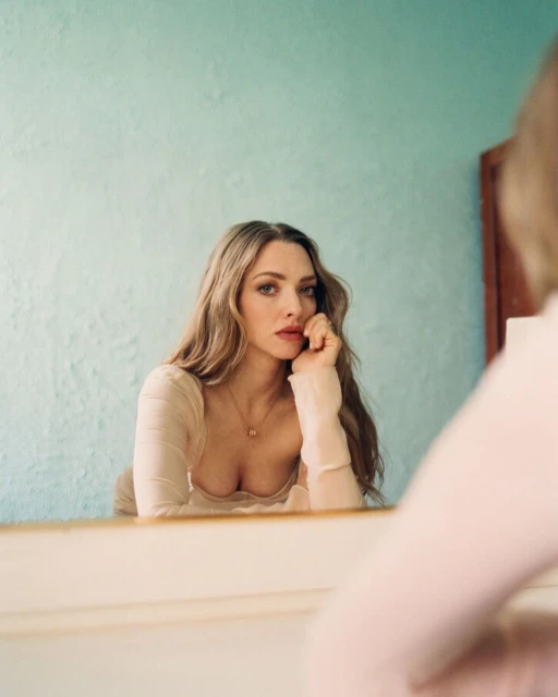 Amanda Seyfried portrait reflected in mirror, wearing a light pink top and necklace, with long blonde hair against a teal wall.