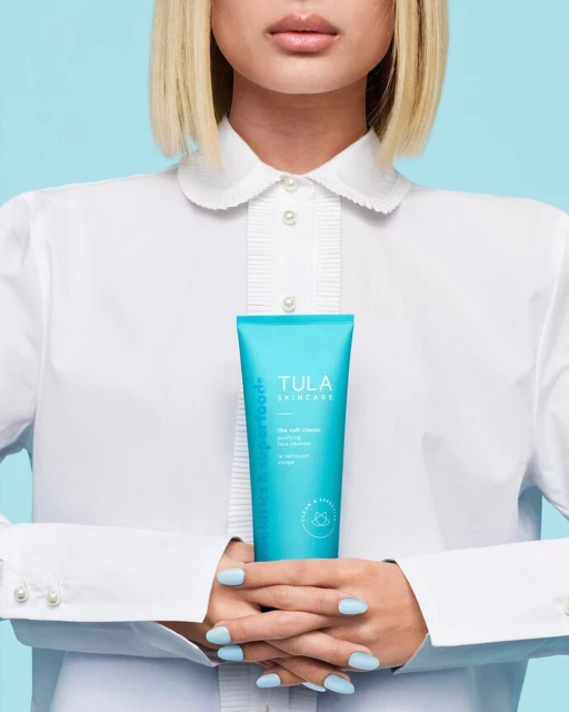 Model showcases Tula Skincare product in a studio shot with a light blue background, wearing a white button-up shirt and holding a turquoise tube.