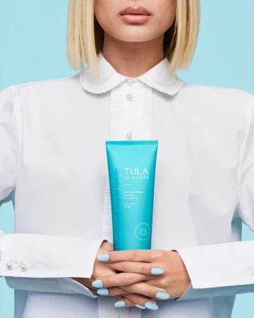 Model showcases Tula Skincare product in a studio shot with a light blue background, wearing a white button-up shirt and holding a turquoise tube.