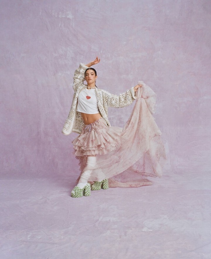 Young woman in motion wearing a heart graphic tee, layered skirt, jacket, and platform shoes poses in front of a textured pink backdrop.