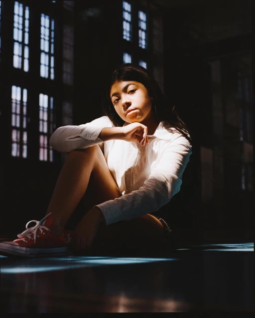 Portrait of a young woman in a white shirt and red sneakers sitting indoors with light streaming through the windows behind her.