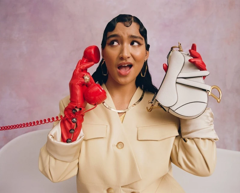 Woman in a cream jacket and red gloves holds a red retro phone and a white purse, looking off to the side against a mottled backdrop.