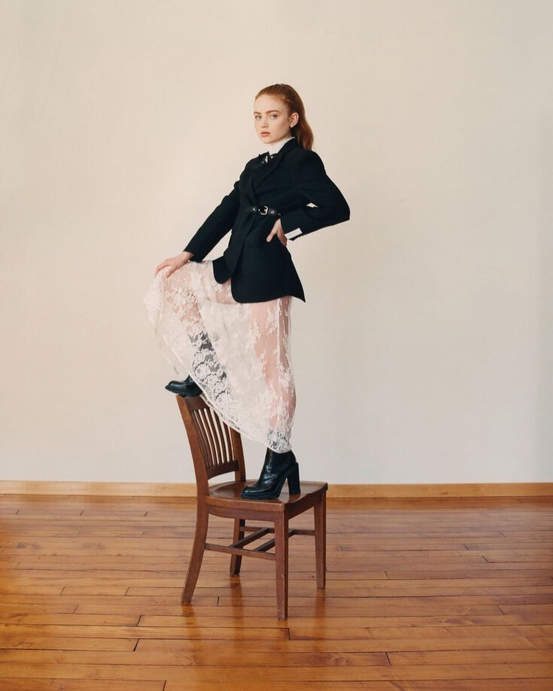A young adult female model posing on a wooden chair in a black blazer and a white lace dress, set against a neutral backdrop in studio.