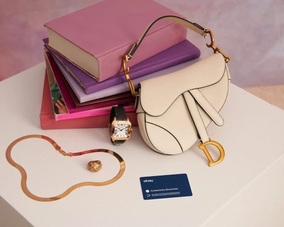Cream-colored Dior saddle bag on a white table with a stack of books, vintage gold jewelry, wristwatch, and an eBay authenticity card.