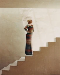 A blonde woman in profile wearing a striped long-sleeve dress stands atop a concrete staircase.