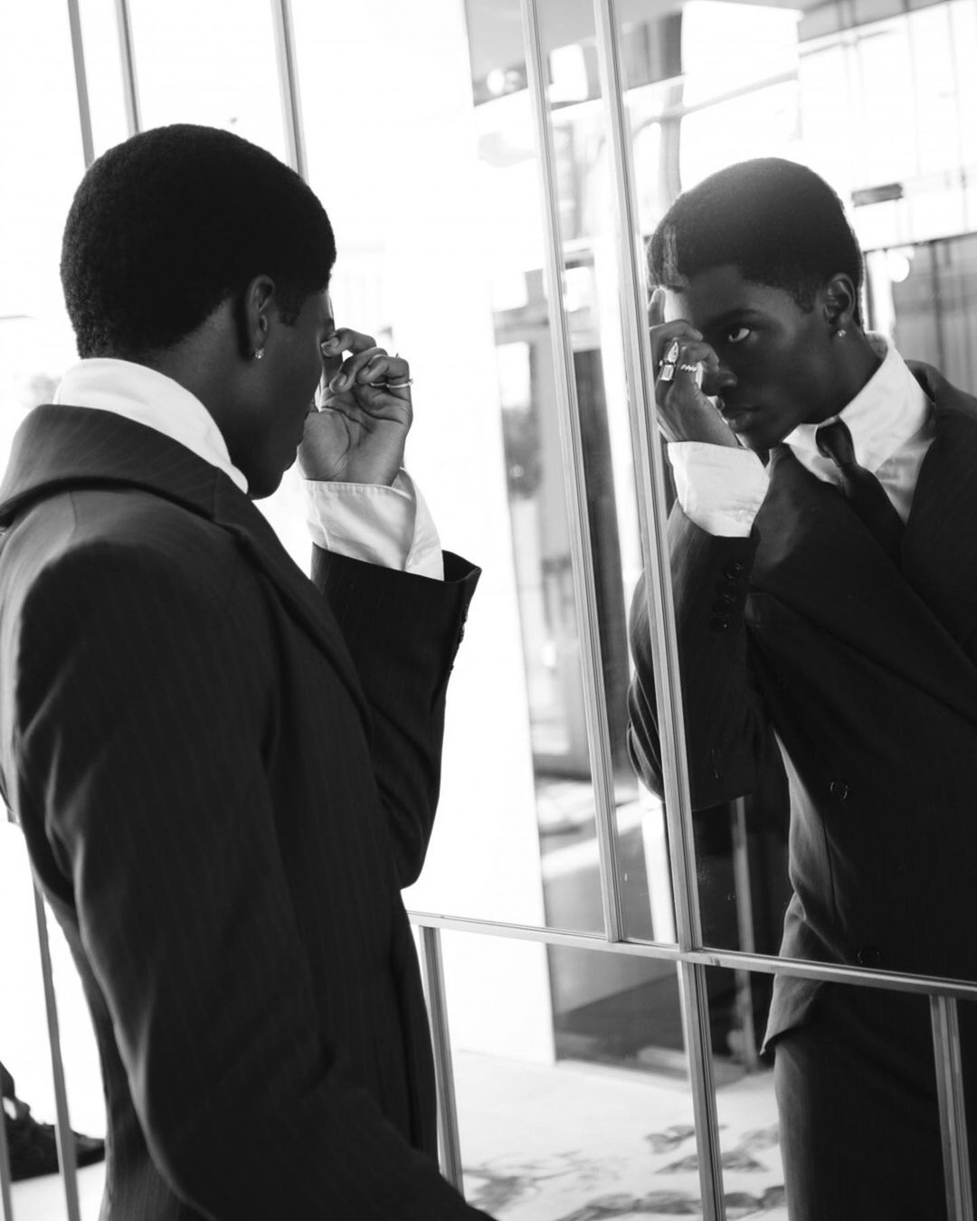A Black man in a suit gazes into a mirror indoors, adjusting his earring, captured in a striking black and white portrait.
