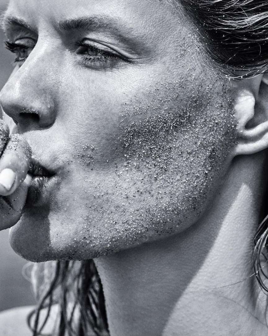 Close-up black and white shot of a woman's face with sand on her cheek and long hair. The image shows a beauty or fashion concept.