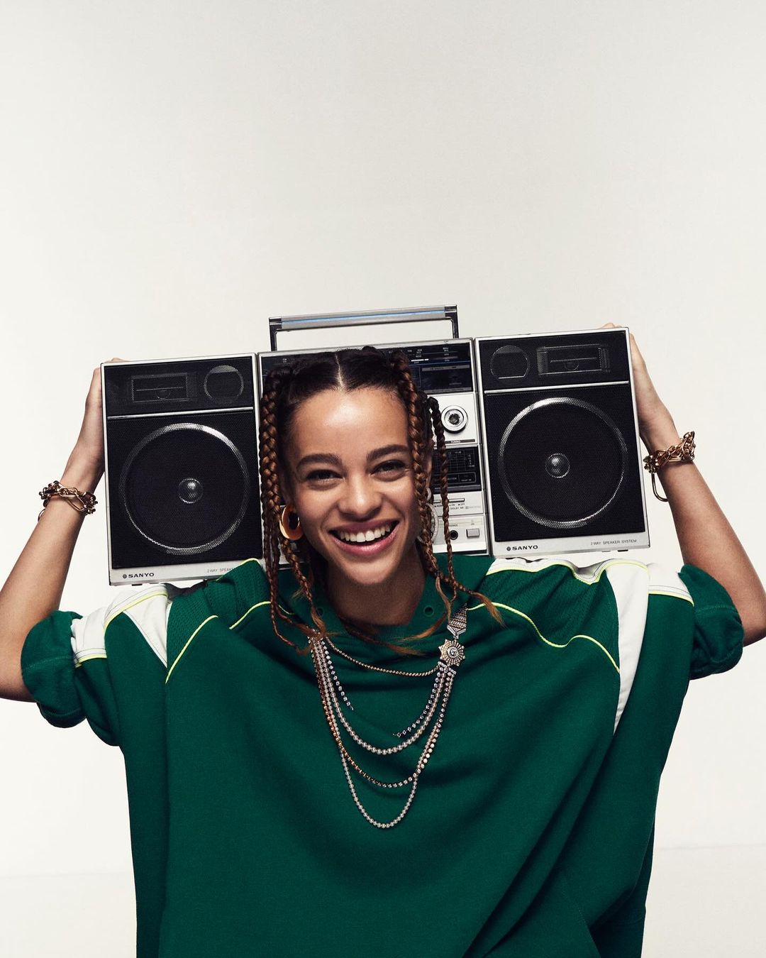 Smiling woman with braided hair wearing a green sweater and jewelry, holding a boombox over her head against a white background.