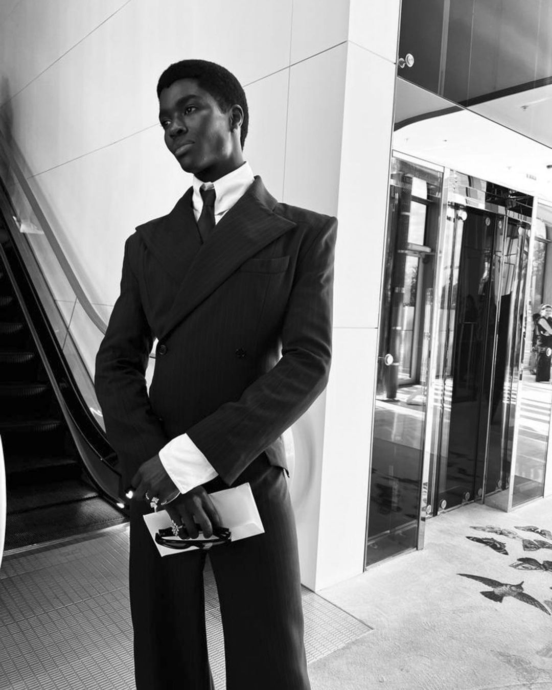 Monochrome shot of a Black male model in a sharp, dark suit. He is holding sunglasses, posing against a modern architectural backdrop indoors.