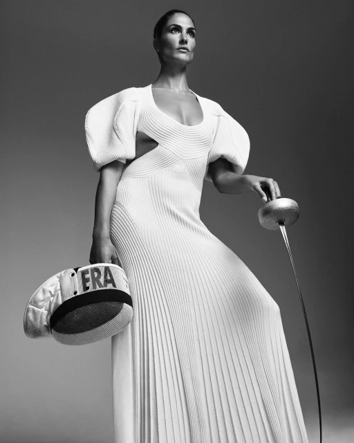 Elegant woman in long white gown holding a fencing foil and mask in a studio setting, creating a striking editorial fashion photograph.